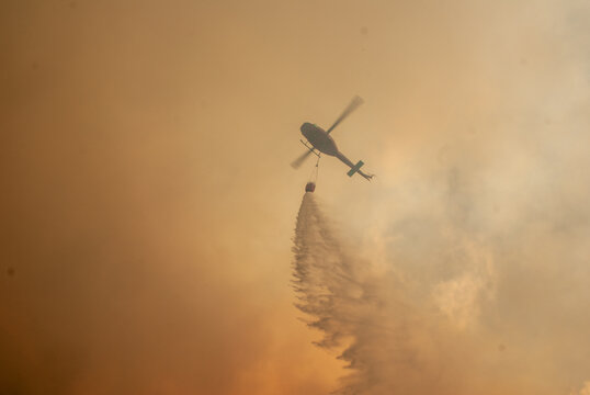 View of a helicopter dropping water on a wildfire with thick smoke filling the sky in South Africa.