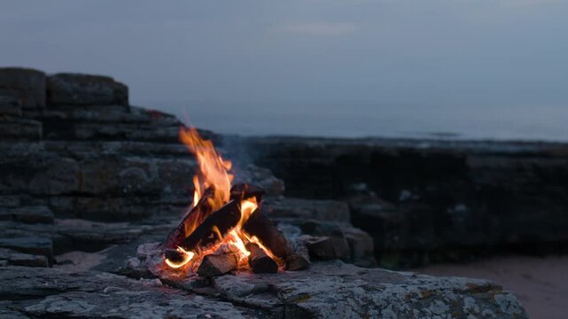 Bright flames flicker and lean in the coastal breeze as a small campfire burns steadily on weathered rocks beside the sea.