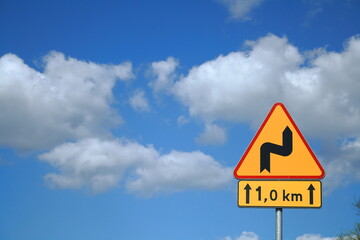 Winding road traffic sign against a blue sky with clouds, warning of upcoming curves. © Wiktor