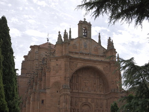 View of the ornate Plateresque facade of the Convento de San Esteban, framed by tall cypress trees under a cloudy sky in Salamanca, Castile and Leon, Spain.