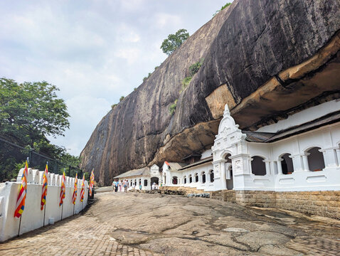View of the historic Dambulla Cave Temple with its white arched facade built into a massive rock face, featuring Buddhist flags along a stone path in Dambulla, Central Province, Sri Lanka.