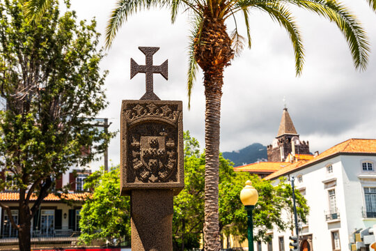 Historical stone monument with the Portuguese cross in sunny Funchal, Madeira, surrounded by palm trees and traditional urban architecture on a bright summer day.