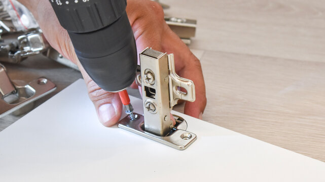 Close-up of a carpenter's hand using an electric drill to screw a metal concealed cabinet hinge onto a white board