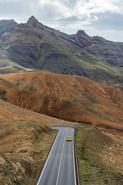 View of a yellow car driving along a winding asphalt road through the rugged, arid volcanic mountains of Fuerteventura in Pajara, Canary Islands, Spain.