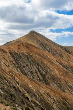 View of the rugged volcanic mountain slopes with textured rock formations under a cloudy blue sky in Pajara, Canary Islands, Spain.
