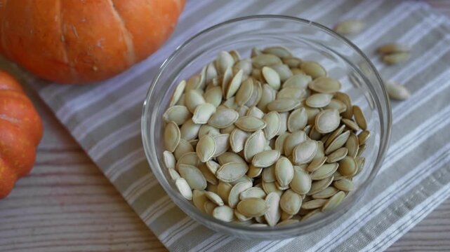 Raw dried pumpkin seeds in glass bowl close up with organic pumpkins on background.