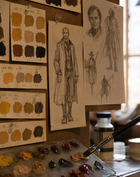 Close up of artist mixing oil paint on glass palette. Character design sketches and color swatches pinned to board in studio. Traditional art and animation pre-production process