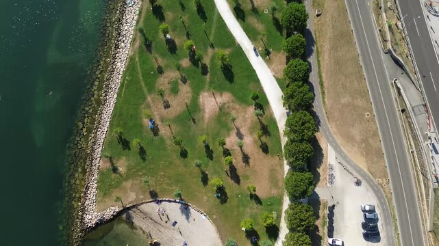 Top down aerial view of the Koper coastline in Slovenia featuring a beach, green hillside, busy coastal road, parked cars and the Adriatic Sea.