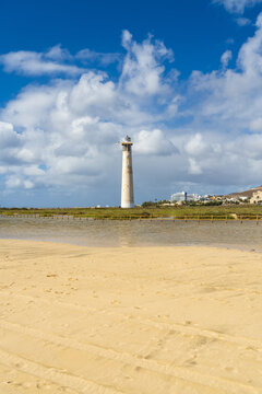 View of the Morro Jable Lighthouse standing on the coast with golden sand in the foreground and a blue sky with clouds in Morro Jable, Canary Islands, Spain.