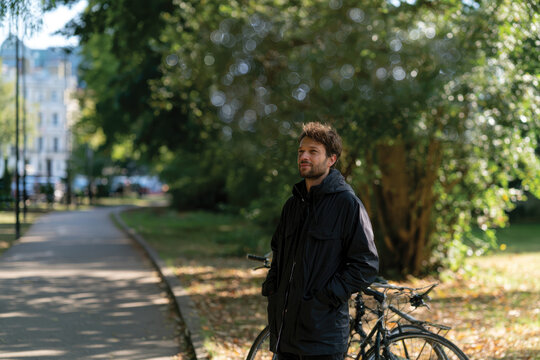 A man stands pensively near his bicycle in a sunlit park, surrounded by trees and nature, capturing a moment of thoughtful introspection amid a vibrant backdrop.