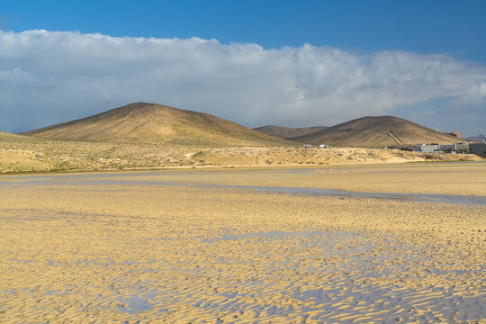 View of the sandy beach at low tide with rolling hills in the background under a blue sky in Jandia, Canarias, Spain.