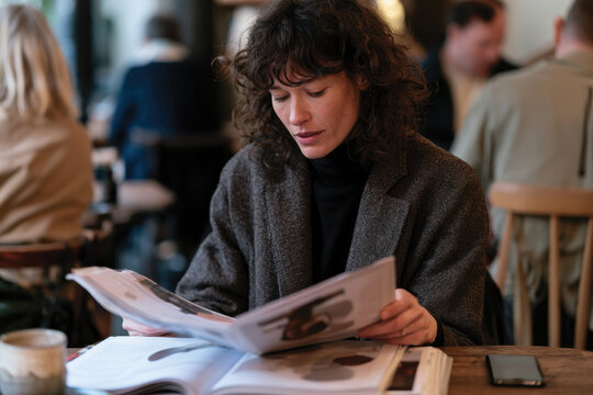 A woman focuses intently on a magazine in a caf&eacute;, capturing a moment of reflection and engagement with visual stories, showcasing modern life in a bustling environment.