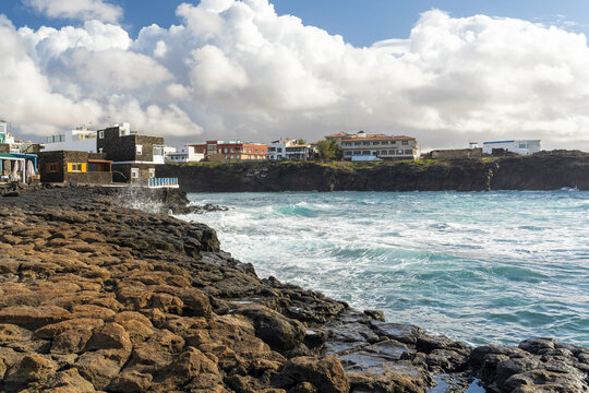 View of the rugged volcanic coastline and turquoise waters of the Atlantic Ocean with white buildings of the village in the background, El Cotillo, Canary Islands, Spain.