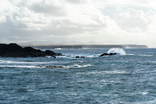 View of waves crashing against the dark volcanic rocks along the rugged coastline under a cloudy sky in El Cotillo, Canary Islands, Spain.