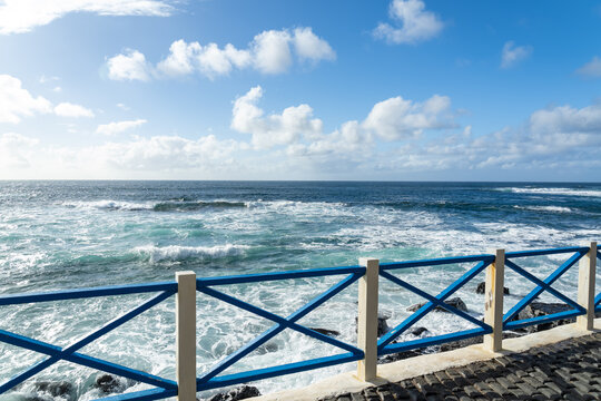 View of the Atlantic Ocean waves crashing against the rocky coastline from a blue wooden fence along a cobblestone path in El Cotillo, Canary Islands, Spain.