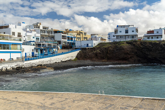 View of the coastal town of El Cotillo with white buildings and blue accents, overlooking a small bay with rocky cliffs under a bright sky in El Cotillo, Canary Islands, Spain.
