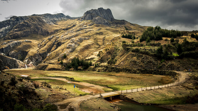 View of Huascaran mountain landscape with a small bridge over a stream in a grassy valley under a cloudy sky in Yungay Province, Ancash, Peru.