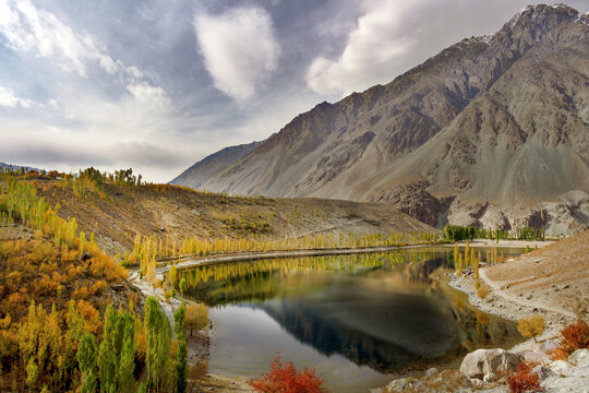 View of Phander Lake surrounded by vibrant autumn trees and towering mountains under a cloudy sky in Phander Valley, Pakistan.
