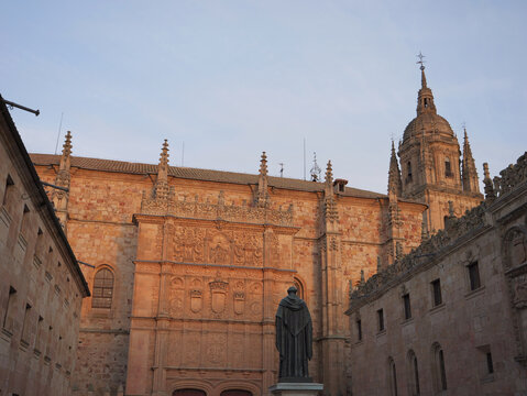 View of the ornate Plateresque facade of the University of Salamanca with a statue in the foreground and the cathedral tower rising against the sky in Salamanca, Castile and Leon, Spain.