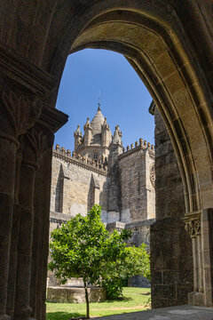 View of the historic Cathedral of Evora framed by a stone archway, featuring Gothic architecture, a small tree in the courtyard, and a clear blue sky in Evora, Distrito de Evora, Portugal.