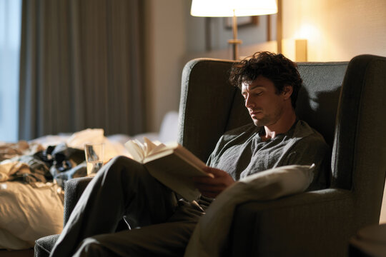 A man sits comfortably in an armchair, immersed in a book, surrounded by a cozy room filled with soft lighting and personal belongings, suggesting relaxation and introspection.