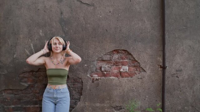 Portrait of Caucasian teenager wearing wireless headphones, listening to music and smiling while leaning against weathered brick wall. Clip is ideal for youth lifestyle and audio accessory commercials