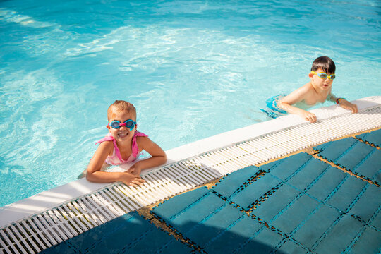 Girl in goggles smiling at pool edge beside boy after summer swimming session. Concept of swim schools, family resorts and children wellness.