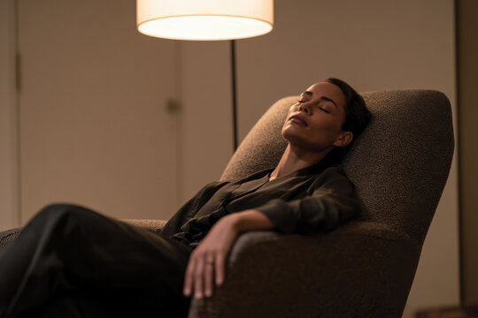 A woman peacefully rests in a chic chair, embracing a tranquil atmosphere, illuminated by soft light, symbolizing relaxation and the art of unwinding in a contemporary space.