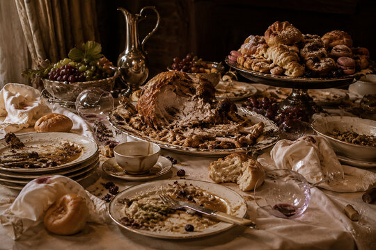 Wide still-life of overloaded banquet table after long meal, torn roast, stacked plates, scattered bread, tipped wine glasses and pastries, gluttony mood