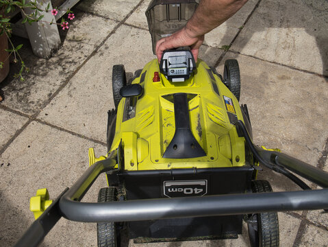 Rear view of a man lifting flap inserting a charged lithium-ion battery into a lawn mower in a backyard