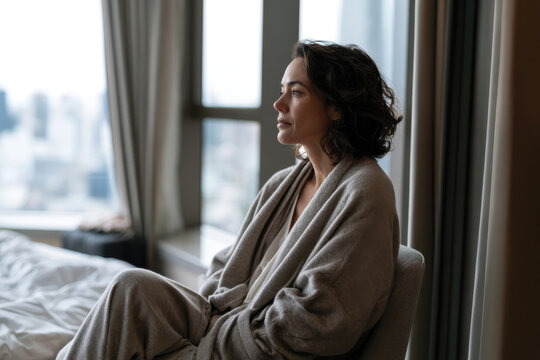 A thoughtful woman wrapped in a cozy robe gazes out of a hotel room window, lost in her reflections while the cityscape provides a calming backdrop during daylight.