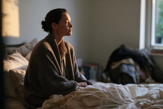 A contemplative woman sits on her bed, engrossed in her thoughts, showcasing a peaceful moment that embodies serenity and introspection within a softly lit room.