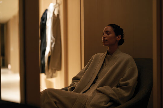 A serene woman, draped in a cozy robe, sits quietly in a softly lit space, exemplifying a moment of relaxation and contemplation in a modern and stylish setting.