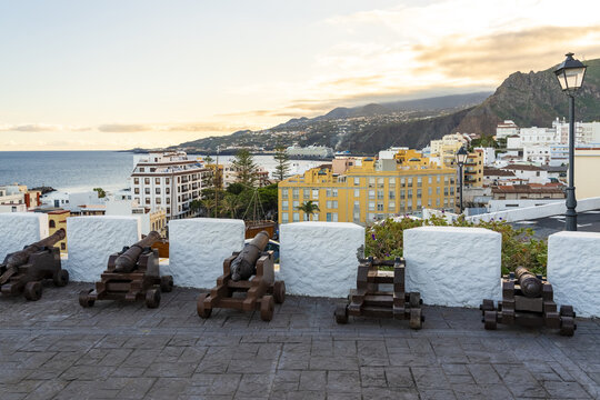 View of historic cannons lined up on a stone wall overlooking the coastal city, harbor, and mountains under a warm sunset sky in Santa Cruz de La Palma, Canary Islands, Spain.