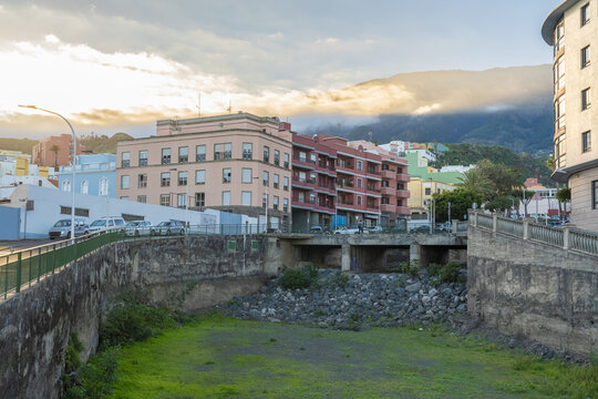 View of colorful buildings, a stone bridge over a dry riverbed, and mountains in the background under a soft sunset sky in Santa Cruz de La Palma, Canary Islands, Spain.