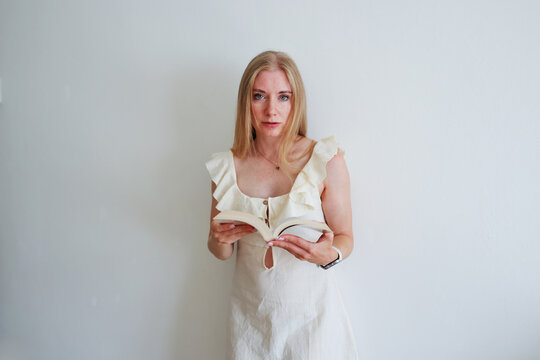 Young blonde woman holding an open book and looking at camera against a white wall with copy space.