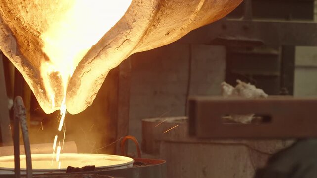 Molten white hot steel flows from a heavy ladle into a round mold as sparks rise in a hot factory bay.