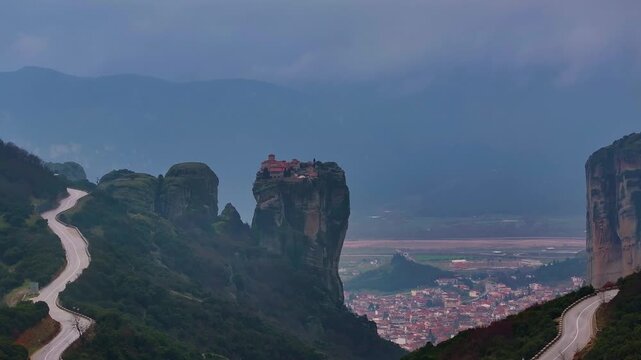 Wide telephoto shot of Holy Trinity Monastery atop a rock pillar with winding mountain roads and Kalambaka town in the valley under moody sky in Meteora, Thessaly, Greece.