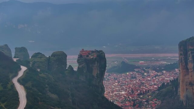 Drone aerial shot rising slowly up, showing Holy Trinity Monastery on a cliff rock, winding mountain road, and Kalambaka town in Thessaly valley under misty overcast sky in Meteora, Greece.
