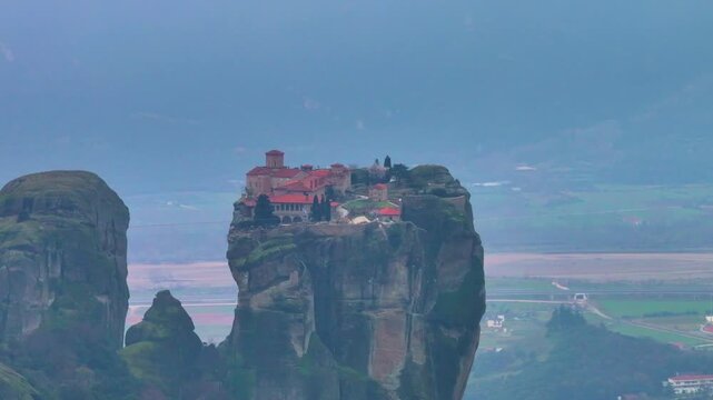 Telephoto shot of Agia Triada Holy Trinity Orthodox Monastery perched on top of a cliff rock in Meteora with Thessaly valley and misty mountains in background under moody overcast light in Greece