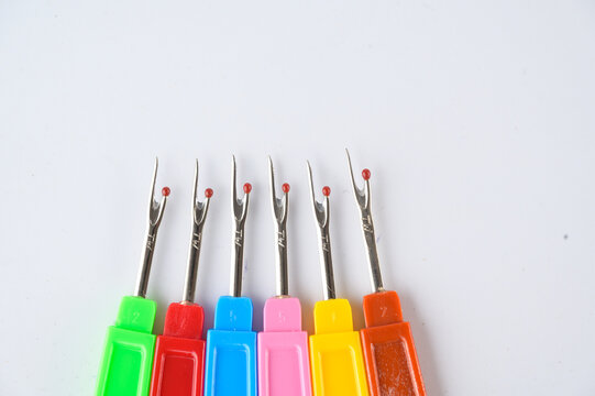 Six colorful seam rippers with metal hooks are aligned horizontally on a white background.