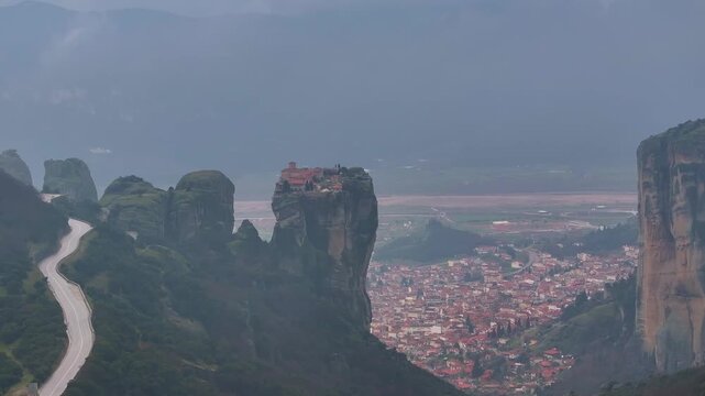 Drone aerial shot slowly panning towards right Holy Trinity Monastery on a rock cliff, with winding mountain road and Kalambaka town in Thessaly valley under misty sky in Meteora, Greece.