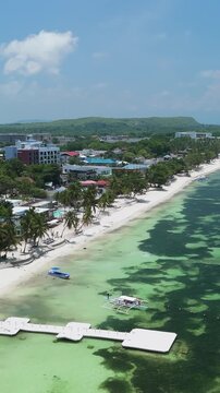 Vertical drone flies over and orbits left above Alona Beach, showing white sand, shallow reef water, boats, and the Panglao coast in Bohol