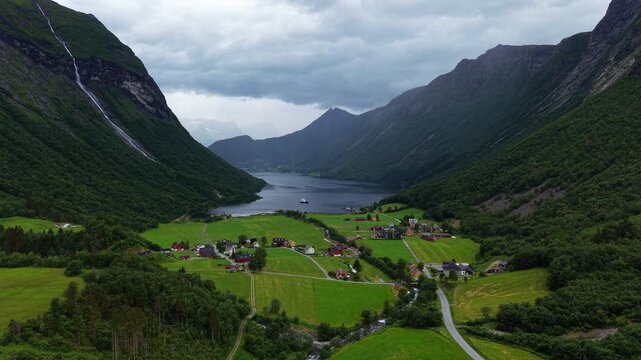 Small fjordside village, bright fields and steep valley walls sit beneath thick summer clouds as a ferry moves along the water.