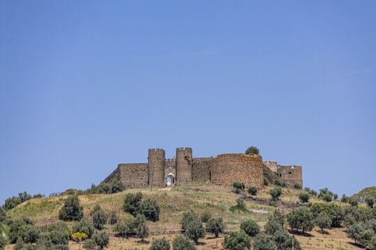 View of the historic stone walls and towers of the Castle of Evoramonte set atop a grassy hill with scattered olive trees under a clear blue sky in Evoramonte, Evora District, Portugal.