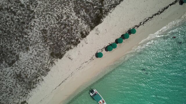 top-down aerial drone shot row of green sun umbrellas lined up on a white sandy beach next to clear turquoise water with a small boat anchored near the shore.