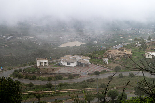 View of the Fort Munro Road winding through the misty hills of the Sulaiman Range, featuring a prominent building complex and a small pond in the distance in Fort Monroe, Punjab, Pakistan.