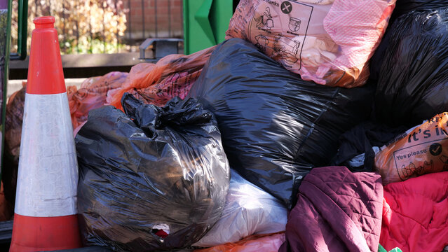 Overflowing rubbish bin area filled with black bin bags recycling sacks and traffic cone outside residential property
