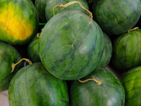 A close-up shot of round, green-striped red watermelons with intact dried stems piled together in a fresh produce display.