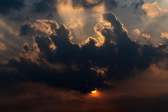 View of the sun setting behind dramatic, dark storm clouds with golden light rays piercing through the sky in Jahanian, Punjab, Pakistan.